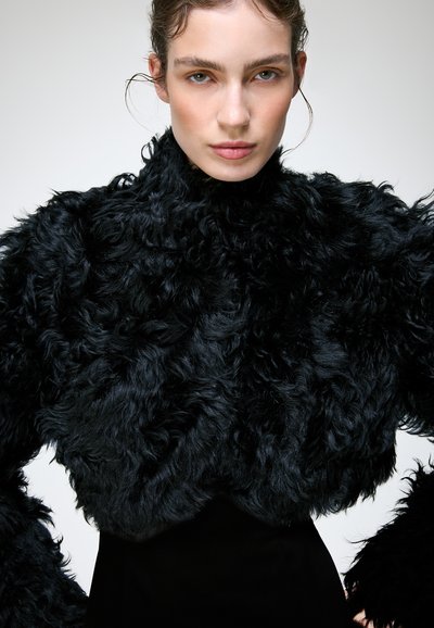 Femme portant une veste volumineuse en fausse fourrure noire avec des boucles texturées, debout devant un fond clair uni.
