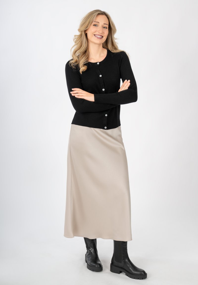 Czarny prążkowany kardigan z okrągłymi guzikami, zestawiony z gładką beżową satynową spódnicą i czarnymi masywnymi botkami za kostkę, stojący na białym tle.