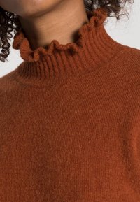 Svetr v rzi barvě s ribovanou texturou a volánkovým límcem. Tkanina vypadá měkce a pohodlně, ideální pro chladné počasí.