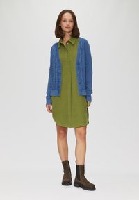Grünes Cordhemdkleid mit Kragen, kombiniert mit einer blauen Strickjacke aus Kabelmuster. Braune Wildleder-Ankle-Boots runden das Outfit ab.