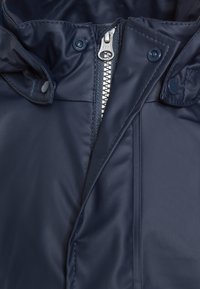 Jachetă puffer bleu închis, cu un material strălucitor, dotată cu un fermoar YKK argintiu, nasturi cu prindere și un guler înalt pentru o căldură suplimentară.
