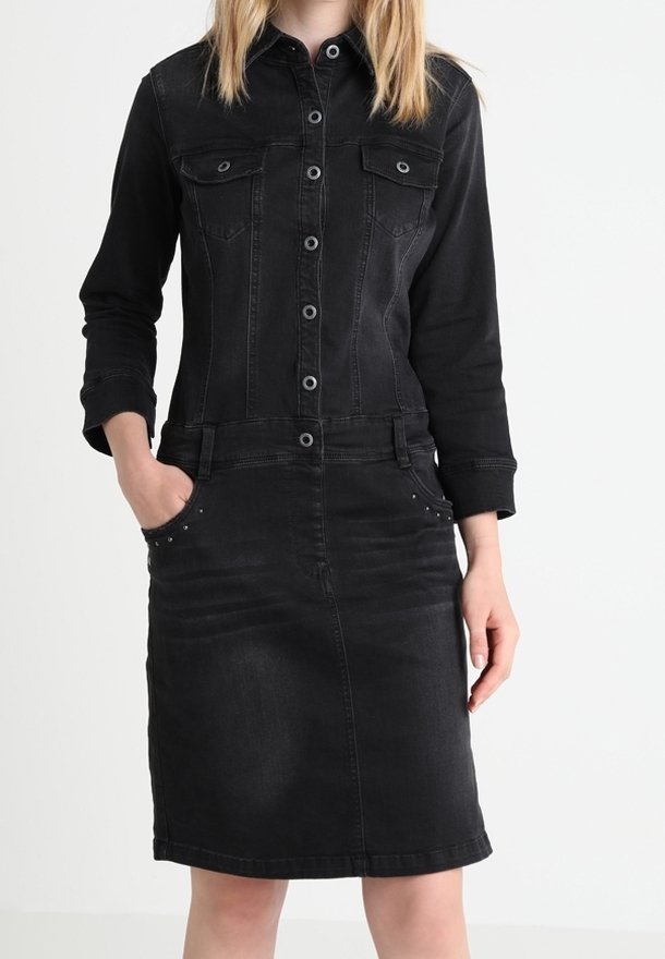 Femme portant une robe en jean noire ajustée avec manches longues, boutonnière devant, poches poitrine et les mains dans les poches des hanches.