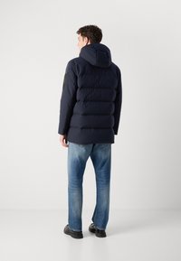 Giacca a vento blu navy con cappuccio, caratterizzata da sezioni a costine orizzontali e una toppa nera sulla manica. Abbinata a jeans azzurri. 