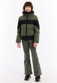 Groene en zwarte pufferjack met een hoge kraag, ritssluiting en zijzakken, gedragen met groene broek en zwarte sneeuwlaarzen.