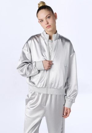 Młoda kobieta z blond kokiem, ubrana w błyszczącą srebrną kurtkę zapinaną na zamek i pasujące spodnie, pozująca na jednolitym białym tle.