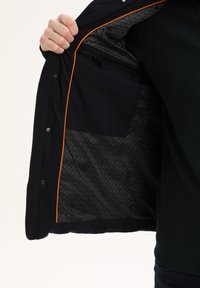 Giacca nera con piping arancione, che mostra un rivestimento interno a fantasia e una tasca con zip, tenuta aperta per rivelare i dettagli del design.
