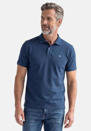 Man met grijs haar en baard, gekleed in een marineblauw poloshirt met korte mouwen en een blauwe spijkerbroek, kijkend iets naar rechts.