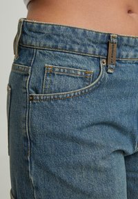 Tuvumā redzams cilvēks, kurš valkā zila denima džinsus, attēlā ir redzams priekšējais kabata, jostas cilpa ar metāla piekariņu un kaila vidukļa daļa virs jostas līnijas.