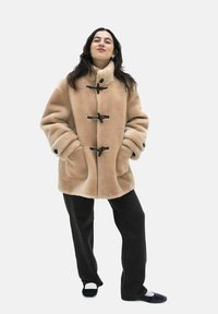 Beige Kunstpelzmantel mit Knopfverschluss, großen Taschen und weiten Ärmeln, kombiniert mit schwarzen Hosen und Schuhen mit einem weißen Akzent.