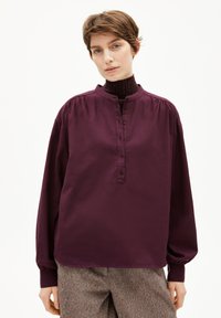 Camicia a maniche lunghe di un profondo viola, con abbottonatura e colletto alto; presenta spalle arricciate e una morbida texture in cotone.