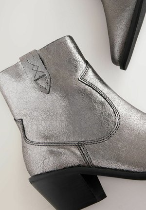 Metalizuoti sidabriniai kulkšniniai batai su tekstūruotu paviršiumi, juodais siūlais ir žemu bloko kulnu. Yra kreivųjų si ūlių dizainas ir logotipo detalė.