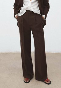 Bruine wijde broek met plooien, gemaakt van gladde stof. Gepaard met een witte gestreepte blouse en zwarte sandalen met rode accenten.