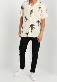 Camisa de manga corta y cuello en color crema con estampado de palmeras negras; combinada con pantalones negros y zapatos blancos. La tela parece ligera.