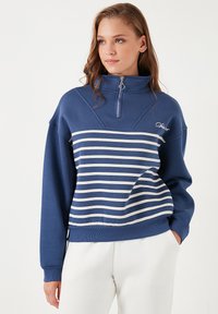 Felpa blu a mezza zip con strisce orizzontali bianche, dotata di un colletto alto e polsini a coste. Dettaglio del logo sul petto. Morbido misto cotone.