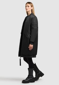 Zwarte gewatteerde jas met een rits aan de voorkant, zijzakken en geribbelde manchetten. Heeft een gestructureerde patch op een van de mouwen en is gecombineerd met zwarte veterboots.