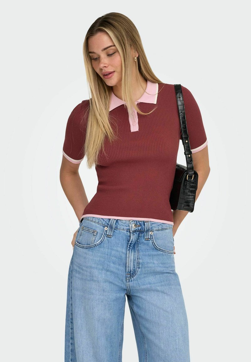 Femme portant un haut côtelé bordeaux avec un col et des bordures roses, un jean taille haute bleu clair et un sac à bandoulière noir, regardant vers le bas.