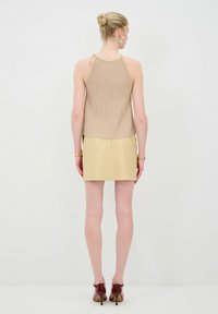 Beige geribbelde top met een gedrapeerd ontwerp en gouden kettingaccenten, gecombineerd met een aansluitende gele leren minirok en bordeauxrode hoge hakken.