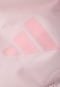 Ροζ αθλητικό ύφασμα με τρία επικάλυπτα, γωνιώδη λογότυπα Adidas σε λίγο πιο σκούρο ροζ. Η επιφάνεια έχει υφή με διάτρητες λεπτομέρειες.