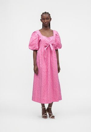Femme grande portant une robe midi rose à manches bouffantes, un nœud devant, et des sandales blanches à talons hauts avec des brides, debout devant un fond uni.