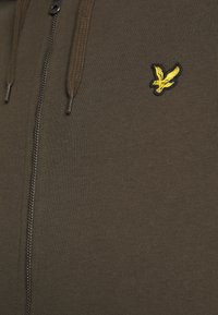 Felpa con zip verde oliva caratterizzata da un tessuto testurizzato, zip metallica e un piccolo logo di un'aquila ricamata gialla sul petto.