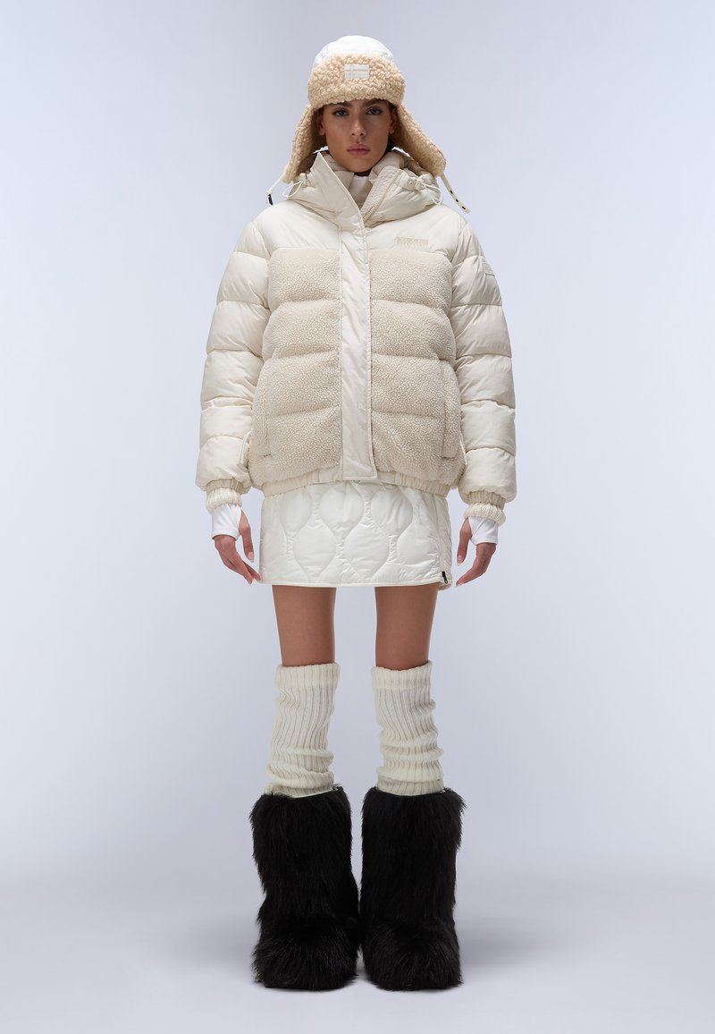 Giacca a vento in crema e beige, con una texture soffice. Presenta una gonna bianca trapuntata, manicotti per le gambe in crema a coste e stivali neri pelosi.