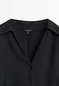 Chemise noire à col avec des boutons sur le devant et une étiquette de taille visible à l'intérieur de l'encolure.