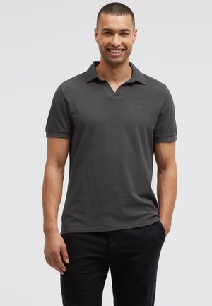 Lächelnder Mann mit kurzen Haaren, der ein dunkelgraues Poloshirt mit kurzen Ärmeln und schwarze Hose trägt, steht mit einer Hand in der Hosentasche.