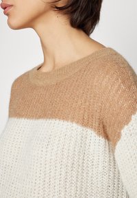 Pull en tricot avec une partie inférieure crème et une partie supérieure beige avec un léger scintillement. Encolure ronde et tissu texturé.