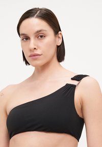 Čierny bikiny top na jedno rameno vyrobený z hladkej látky s nariasenými detailmi a kovovým doplnkom na ramienku.