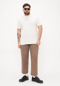 Beige cotton T-shirt with short sleeves and a small graphic, paired with brown straight-leg trousers and black shoes. Accessories include sunglasses.