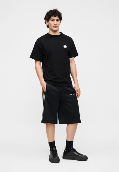 Νέος άνδρας που φοράει μαύρη μπλούζα, μαύρα σορτς με πλαϊνές ρίγες, μαύρες κάλτσες και μαύρα αθλητικά παπούτσια, σε απλό φόντο.