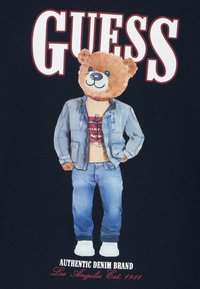 Grafika medveda v denim jakni, majici in modrih kavbojkah, z belimi supergami, na temnem ozadju, ob krepkem napisu "GUESS".