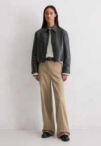 Czarna skórzana kurtka cropped nałożona na paskowaną koszulę z długim rękawem, w połączeniu z beżowymi spodniami w stylu dzwonów i czarnymi butami. Gładka tekstura z guzikami.