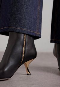 Botte de cheville en cuir noir avec un détail de fermeture éclair dorée et un talon courbé doré, sur fond de denim sombre.
