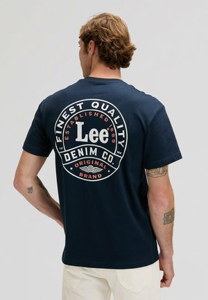 Camiseta de algodón azul marino con un logo circular blanco y rojo que dice "Lee Denim Co." en la parte trasera, con texto gráfico y diseño con alas.