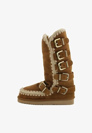 Bota alta de ante marrón con costuras en color crema y cinco hebillas ajustables a lo largo del costado, suela de goma plana y suave forro interior visible en la parte superior.