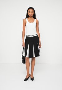 Czarna spódnica z białymi panelami, plisowany design, w połączeniu z białym topem na ramiączkach. Modelka trzyma małą czarną torebkę; ma na sobie czarne szpilki.