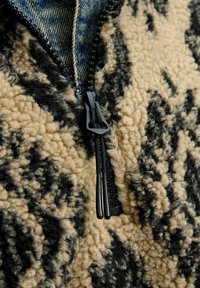 Fleecejacke mit beige Grundfarbe und schwarzen Camouflage-Mustern, hohem Kragen und einem Reißverschluss vorne, der schwarz ist und aus strukturiertem Material besteht.