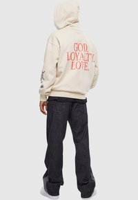 Cremefarvet hættetrøje med rød tekst "GOD. LOYALTY. LOVE." på bagsiden; kombineret med mørke jeans og hvide sneakers. tekstureret stof, afslappet pasform.