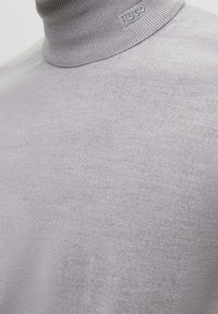 Jasnoszary sweter z golfem wykonany z gładkiej, drobnej dzianiny. Posiada subtelny prążkowany kołnierz i jest oznaczony logo przy linii szyi.