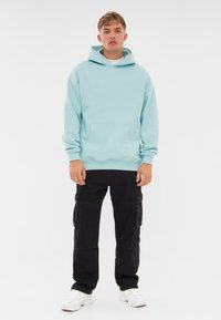 Felpa con cappuccio blu chiaro, con tasca a marsupio, abbinata a pantaloni cargo neri con più tasche laterali e sneakers bianche.