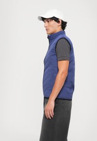 Jovem com um boné branco, colete azul acolchoado, camisa preta de manga curta e jeans pretos desbotados, posando de lado contra um fundo branco.