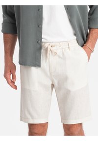 Shorts beige clair avec une taille élastique et un cordon de serrage, dotés d'une finition texturée et d'un ourlet replié. Portés avec une chemise grise à boutons.
