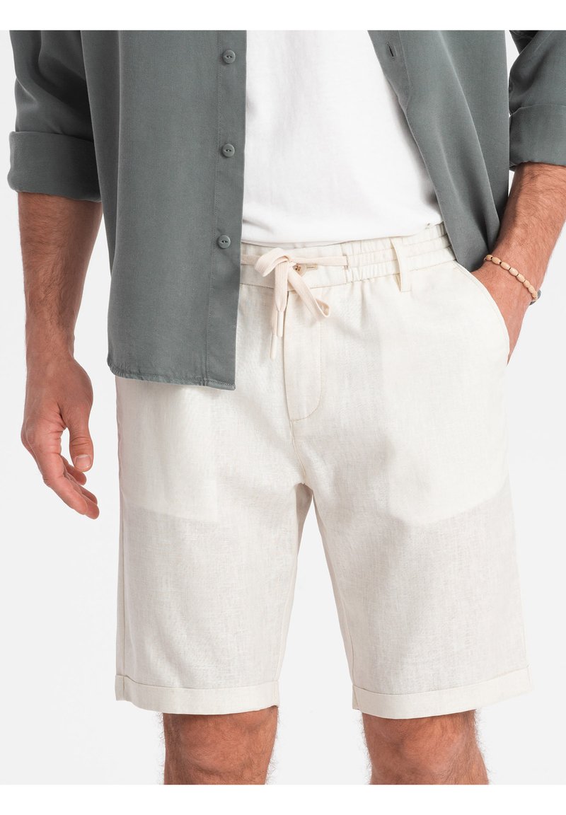 Shorts beige clair avec une taille élastique et un cordon de serrage, dotés d'une finition texturée et d'un ourlet replié. Portés avec une chemise grise à boutons.