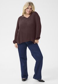 Brown long-sleeve blouse with a V-neckline, relaxed fit, and slightly curved hem, paired with blue straight-leg jeans and black flats.