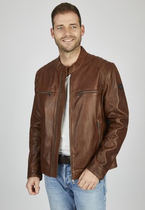 Chaqueta de cuero marrón con cuello alzado, dos bolsillos en el pecho con cremallera y detalles de costura horizontal en los hombros. Lleva puesta una camiseta gris.