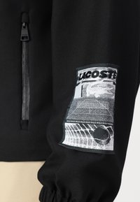 Black jacket featuring a textured fabric, with a prominent graphic patch on the sleeve depicting a tennis court and the word "LACOSTE."
