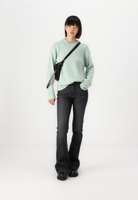 Pull en maille côtelée vert clair, sac bandoulière noir, jean évasé foncé et chaussures noires. Design simple avec des accents et textures minimaux.