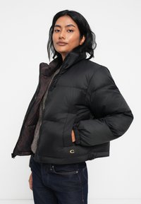 Veste puffer noire avec un col haut, présentant une doublure intérieure à motif, une fermeture éclair et des poignets élastiques. Détail du logo doré sur l'ourlet.