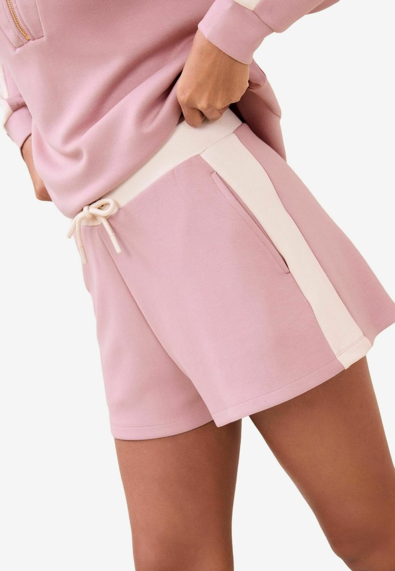 Osoba w jasnoróżowych szortach z białymi paskami po bokach i pasującej różowej bluzie, ręce spoczywają na talii, na białym tle.